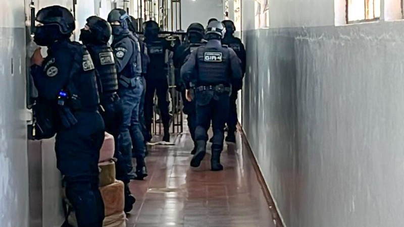 Foto colorida mostra vários operacionais do Grupo de Intervenção Rápida da 4ª Região no corredor da Penitenciária, parede cinza, em pé, de perfil e de costas, vestidos de preto e com capacetes de proteção, vistoriando celas pela janelinha de comunicação.