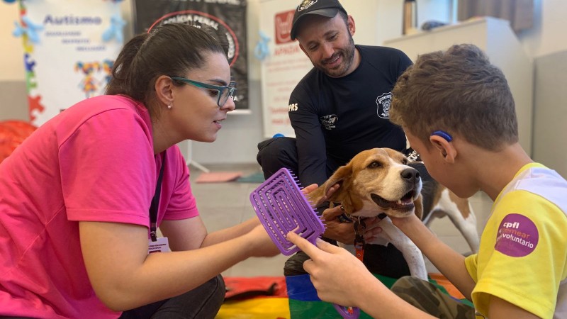 Na imagem, uma mulher de camisa rosa auxilia um menino na terapia com um cachorro. Um policial penal ao fundo, vestido de preto, observa a ação.