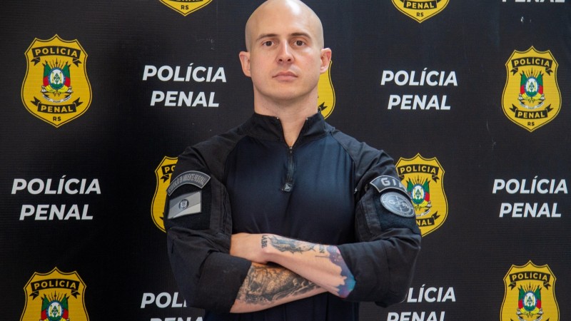Um home branco vestido com a farda da Polícia Penal e de braços cruzados olha seriamente para a camera. Ao fundo, um banner escrito Polícia Penal