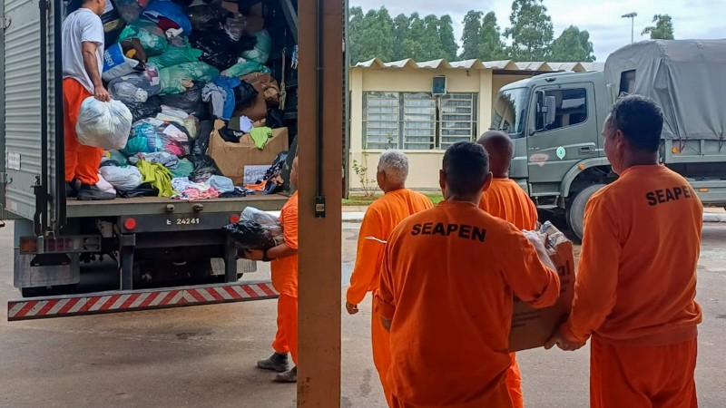 Na foto, homens de vestimentas laranjas estão carregando doações na frente de um caminhão cheio de sacolas com roupas. Dentro do veículo, um homem com camiseta branca e calça laranja segura uma sacola