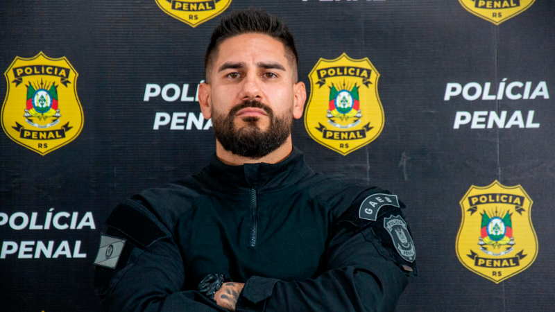 Policial fardado de braços cruzados em frente a banner da Polícia Penal