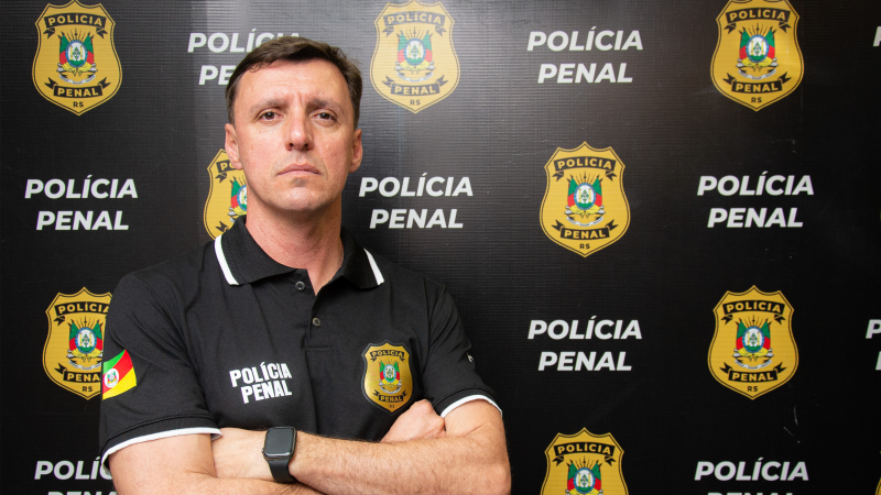 Homem de camisa preta de braços cruzados em frente a banner da Polícia Penal