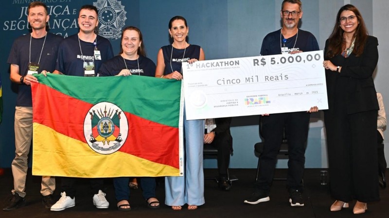 Equipe composta por uma policial penal, professores e um aluno da pós-graduação da Universidade de Santa Cruz do Sul segurando a bandeira do RS recebem premiação